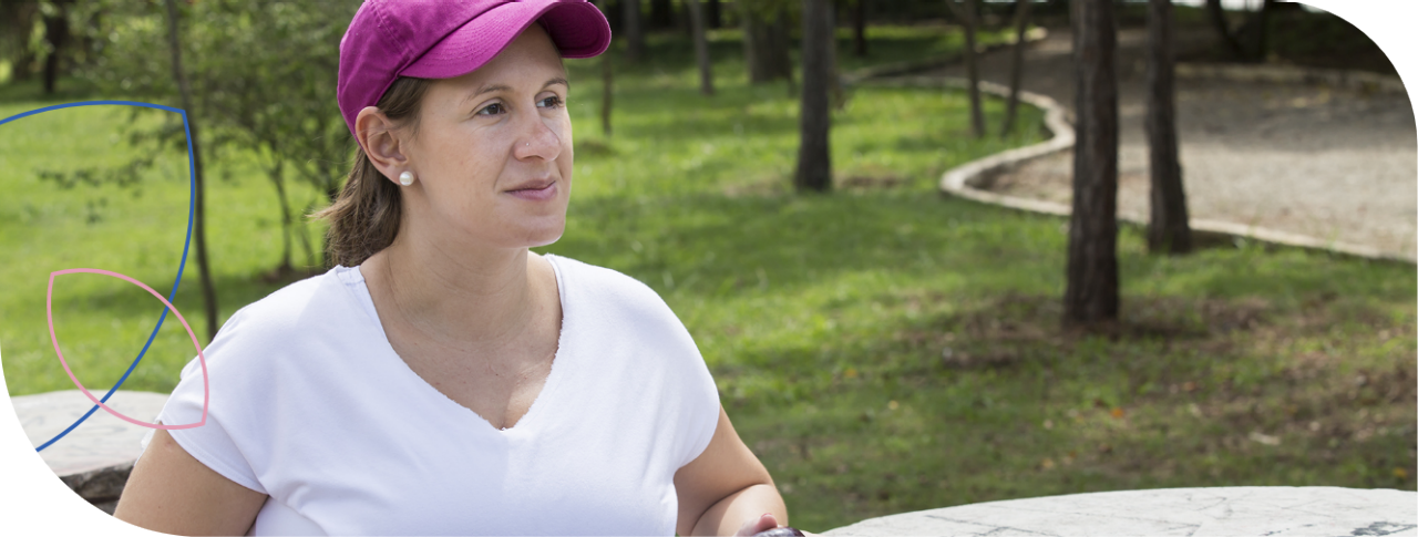 Mulher branca, vestindo blusa branca e boné roxo, sentada em um parque