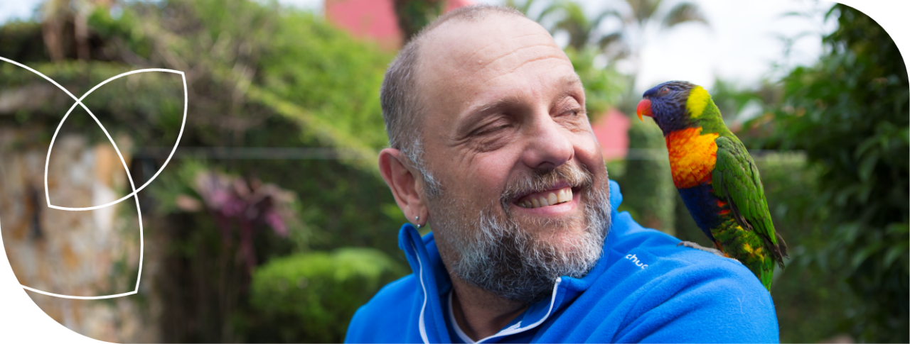 Homem branco sorrindo, com barba e cabelo grisalhos, vestindo camisa azul, com um pássaro Loriini sobre o ombro esquerdo, portador de diabetes tipo 2