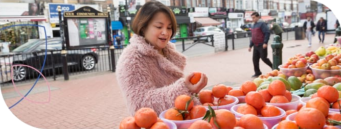 Mulher asiática, cabelos curtos e escuros, veste casaco cor salmão, na feira escolhendo tangerina e outras frutas para sua dieta balanceada de controle da diabetes