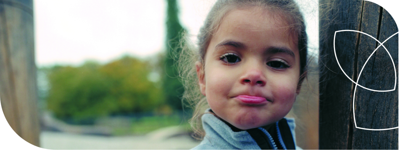 criança de cabelos presos usando casaco com zíper, em close-up, ao ar livre próximo a um tronco de madeira.