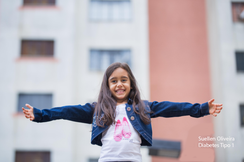 Suellen Oliveira é uma criança portadora de Diabetes Tipo 1, cabelos longos e escuros, veste blusa branca e jaqueta jeans azul, sorrindo e com os braços abertos