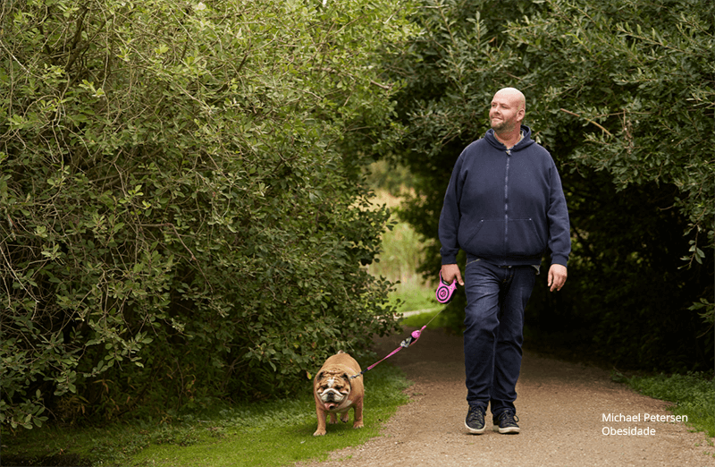 Michael Petersen, paciente obeso, usa calça jeans e moletom escuros. Passeando com seu buldogue francês em um parque com vegetatação densa