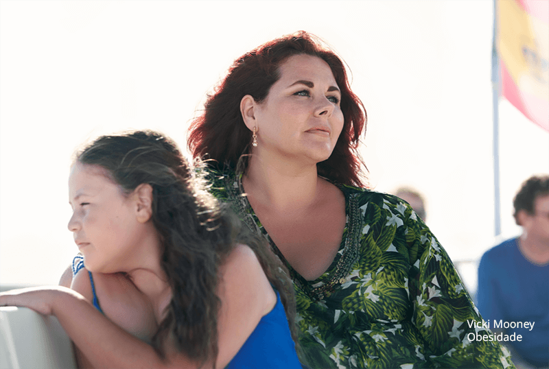 Mãe e filha apresentando sinais de obesidade. Pessoas brancas, a criança usa blusa azul, a mãe blusa com estampas verdes, sentadas em um barco olhando a paisagem