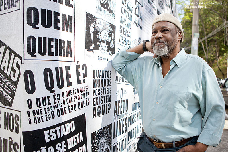 &quot;  Otavio Domingos da Costa, tem diabetes tipo 2, é uma pessoa preta com barba grisalha. Usa camisa verde claro e boina cor nude, apoia o braço em uma parede na rua e esboça sorriso&quot;