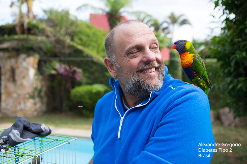 Alexandre Gregório tem diabetes tipo 2, é um homem branco com barba e cabelos grisalhos, veste camisa azul e pousa com um pássaro Loriini sobre o ombro esquerdo